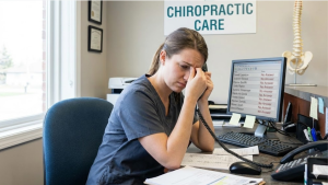 A stressed chiropractic assistant in scrubs sits at a front desk, holding a phone and rubbing her face in frustration. Her computer screen displays a list of patient names with "No Answer" written in red next to each entry. A "Chiropractic Care" sign and a spine model are visible in the background.